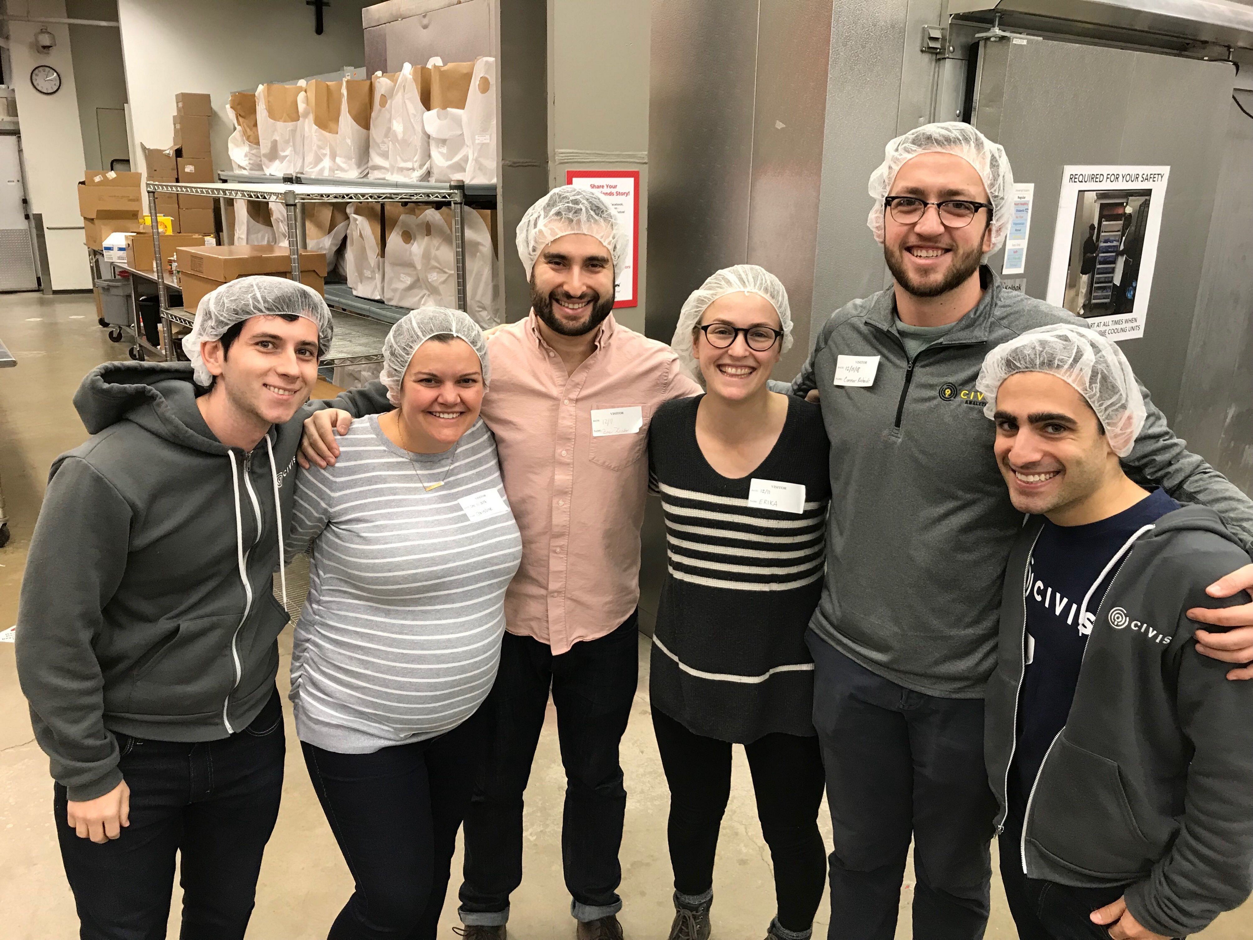 Six employees in hairnets standing in a commercial kitchen 