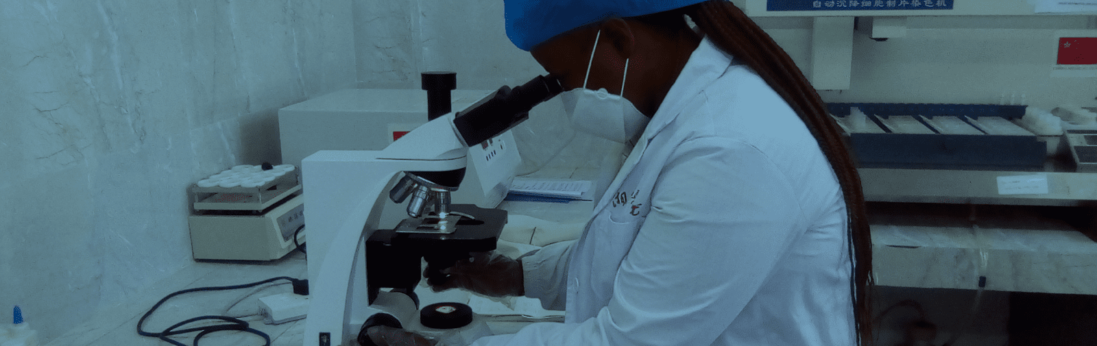 Photo of a woman in a face mask and lab coat looking at a slide under a microscope